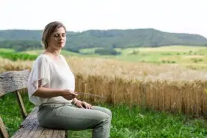 Angela Hügli sitzt mit geschlossenen Augen auf einer Holzbank am Feldrand und hält eine Einhandrute – in einem Moment der Stille, verbunden mit sich und der Natur.