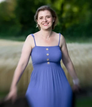 Angela Hügli lächelnd in einem violettblauen Kleid vor einem Baumfeld