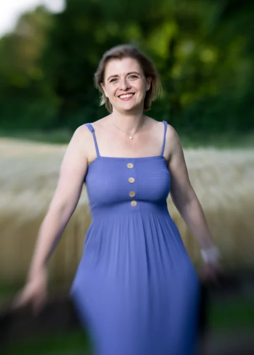 Angela Hügli lächelnd in einem violettblauen Kleid vor einem Baumfeld